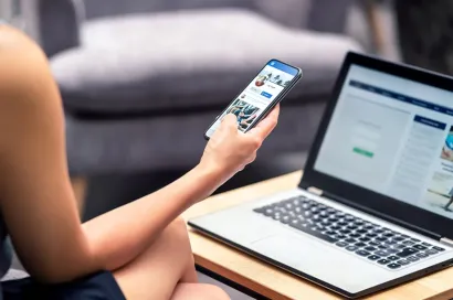 Person sitting with a laptop on a table and a smartphone in hand, browsing Facebook.